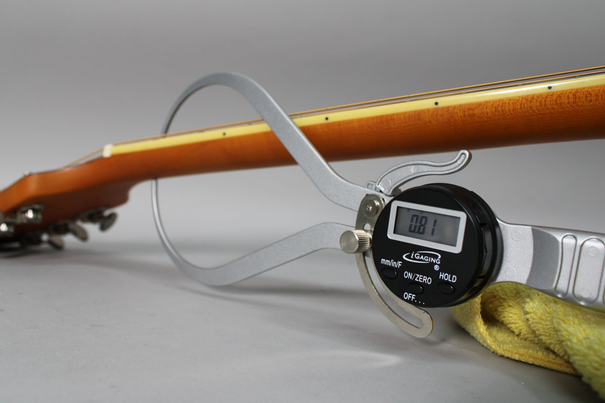 The guitar's natural matte finish neck is measured by a digital caliper, showing a 0.81 reading, with tuning pegs visible.