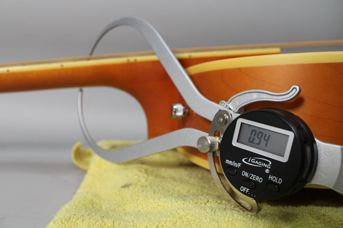 A digital caliper measures the guitar's neck thickness at 0.94, resting on a yellow cloth against the natural matte body.