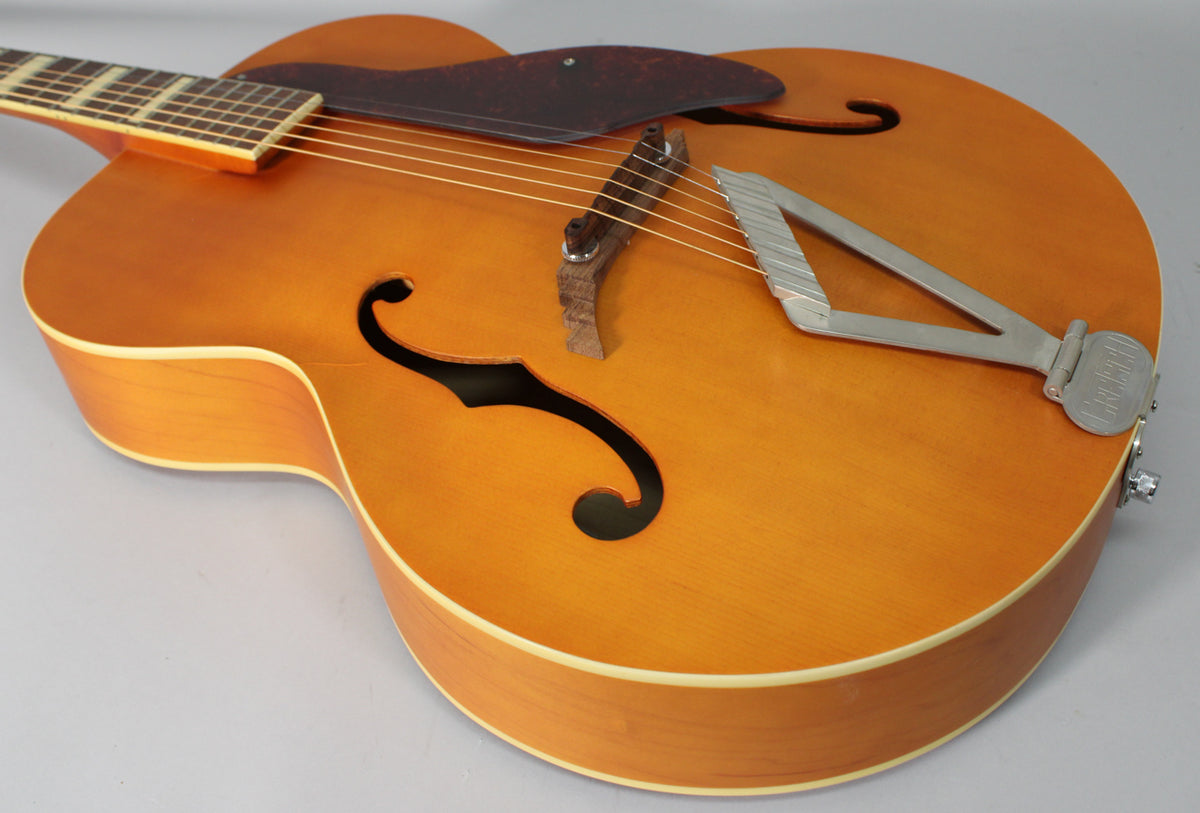 The natural matte archtop guitar is angled from the upper left, showing the f-holes, pickguard, wooden bridge, and tailpiece.