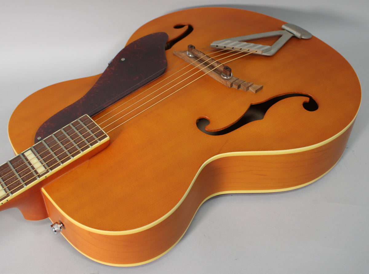 Upper body of a natural matte archtop guitar, showing the f-holes, dark pickguard, and a silver trapeze tailpiece.