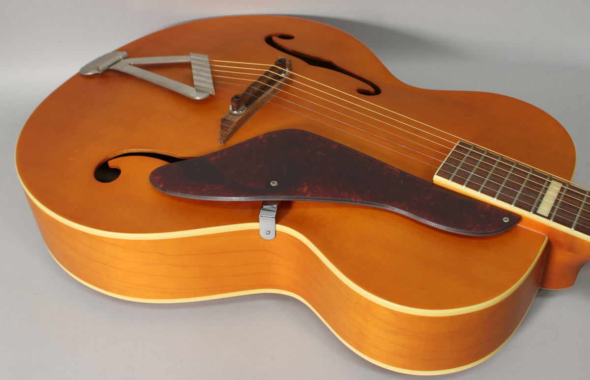 The natural matte archtop guitar is angled from above, showing the body, f-holes, tortoise pickguard, and tailpiece.