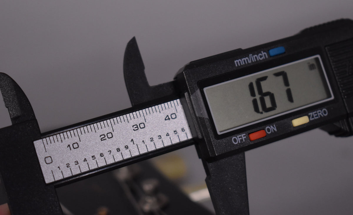A digital caliper measures the nut width at 1.67 inches, with the guitar's headstock and tuning pegs blurred in the background.