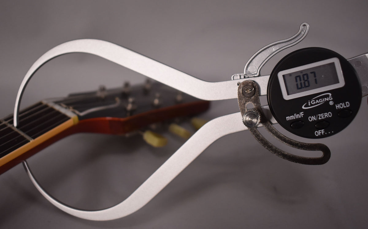 A digital caliper measures the guitar's neck thickness at 0.87, with the headstock and tuners visible in the background.