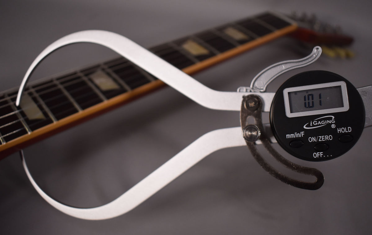 A digital caliper measures the guitar's neck thickness at the first fret, showing "1.01" on its screen, with trapezoid inlays visible.