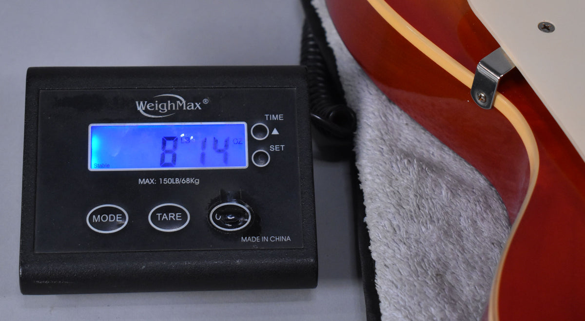 The guitar's cherry sunburst body and cream binding rest on a grey towel, with a scale displaying "8lb 14oz" in the foreground.