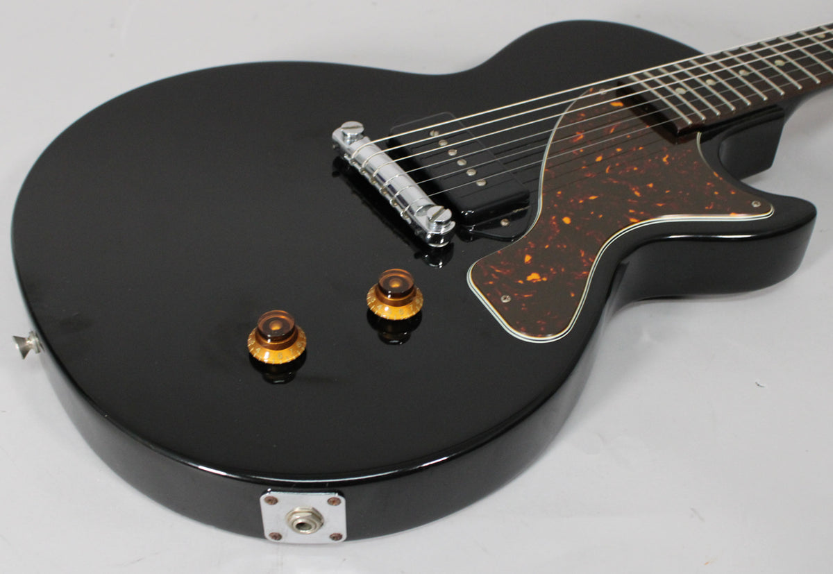The glossy black guitar body is angled, showing the chrome bridge, black P90 pickup, tortoiseshell pickguard, and two amber knobs.