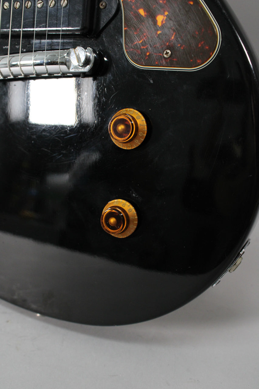 Close-up of the glossy black body, showing two amber "top hat" control knobs, a tortoiseshell pickguard, and light surface scratches.