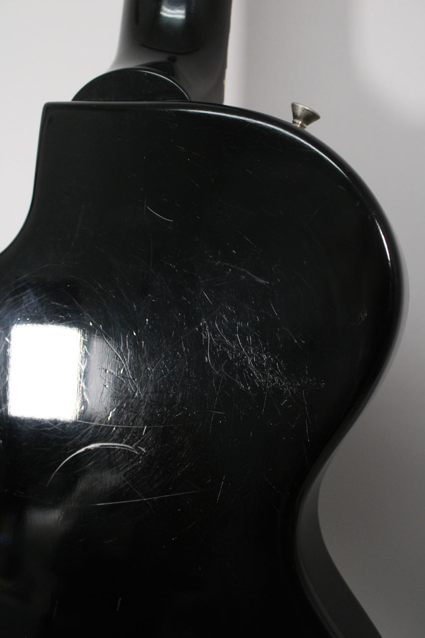 The guitar's glossy black body, viewed from the back, shows extensive surface scratching and a silver strap button on the upper horn.