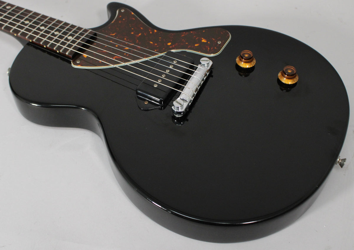 Angled view of the glossy black body, showing the tortoise pickguard, single P90 pickup, chrome bridge, and two amber knobs.