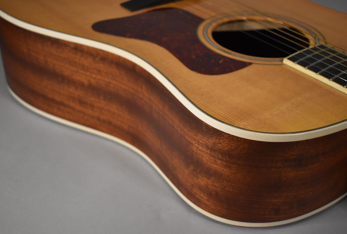 The natural finish acoustic guitar's lower bout and side are framed, showing a dark brown wood grain and cream binding.