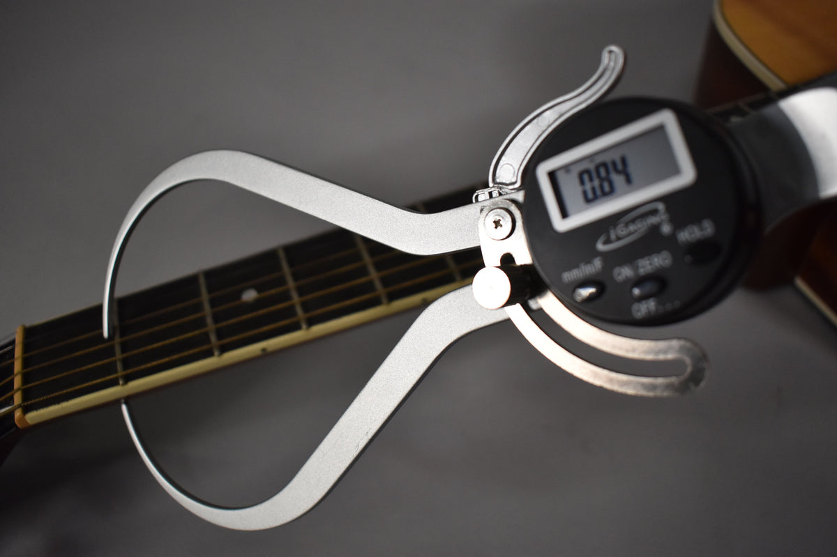 A digital caliper measures the natural finish dreadnought's fretboard, displaying "0.84" on its screen.