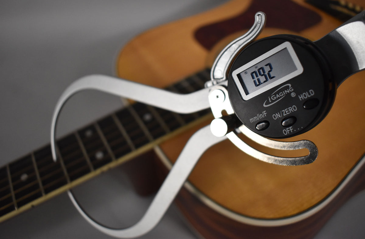 A digital caliper measures the fret height at 0.92mm on the natural finish acoustic guitar's dark fretboard, with its light wood body blurred behind.