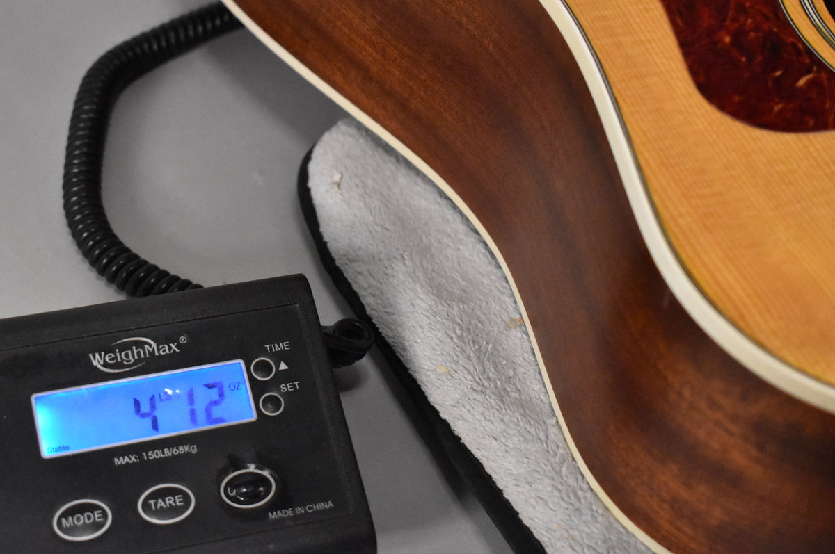 The guitar's natural finish body is shown on a scale displaying 4 lbs 12 oz, with a plush-lined case visible.