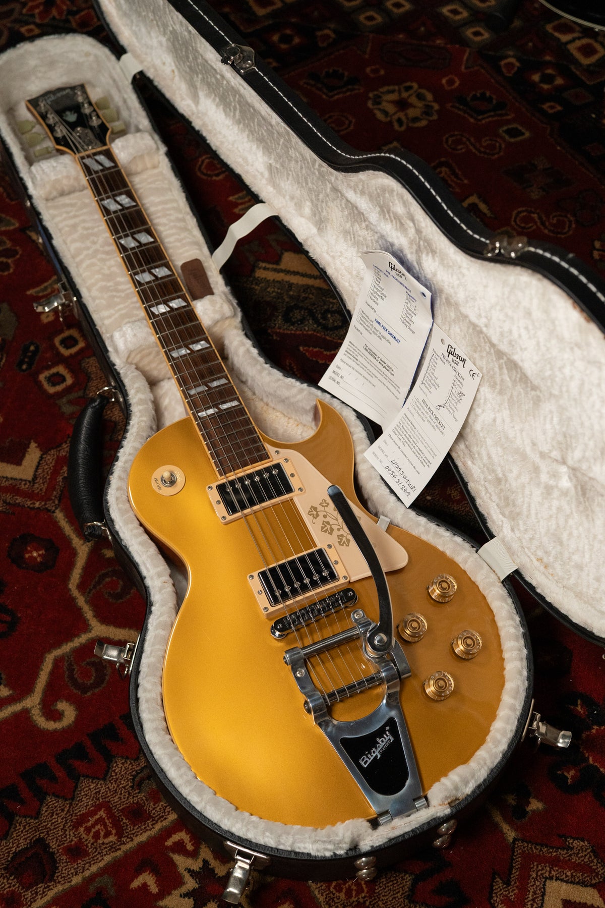 Goldtop Les Paul in its open hard case, angled from above, showing the Bigsby tremolo, etched pickguard, and paperwork.
