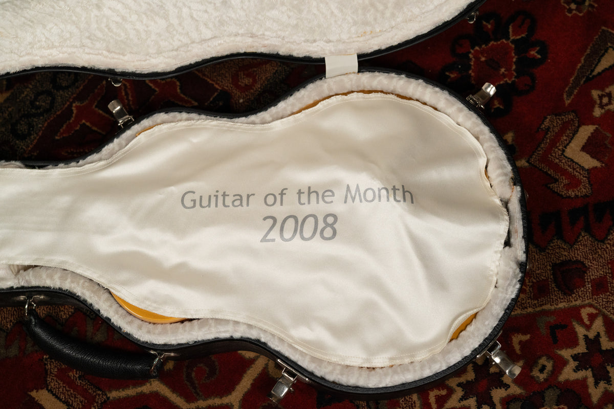 Overhead view of an open black hard case with white plush lining, a white silk shroud with "Guitar of the Month 2008" printed on it.
