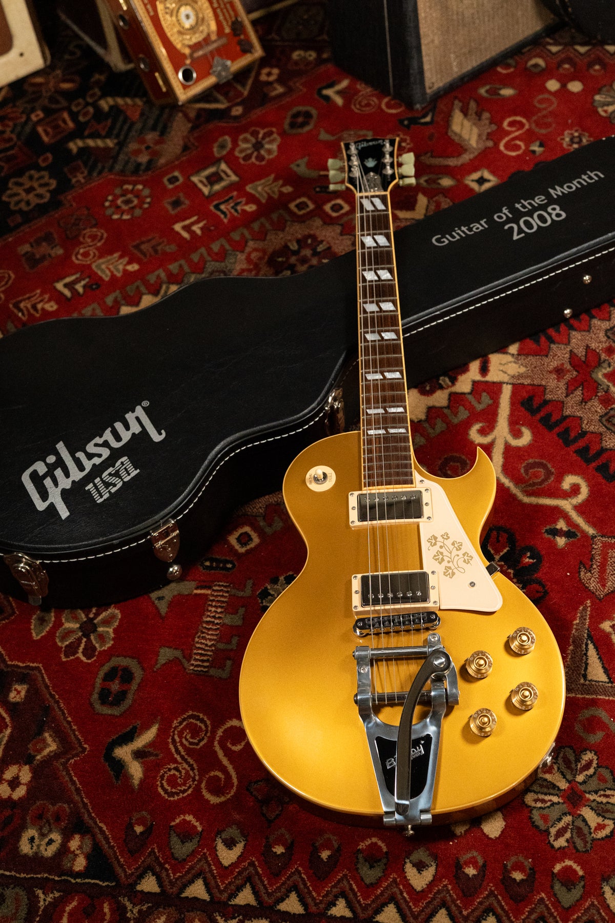 A goldtop Les Paul with a Bigsby vibrato, white pickguard with a shamrock design, and a black hard case, on a red rug.