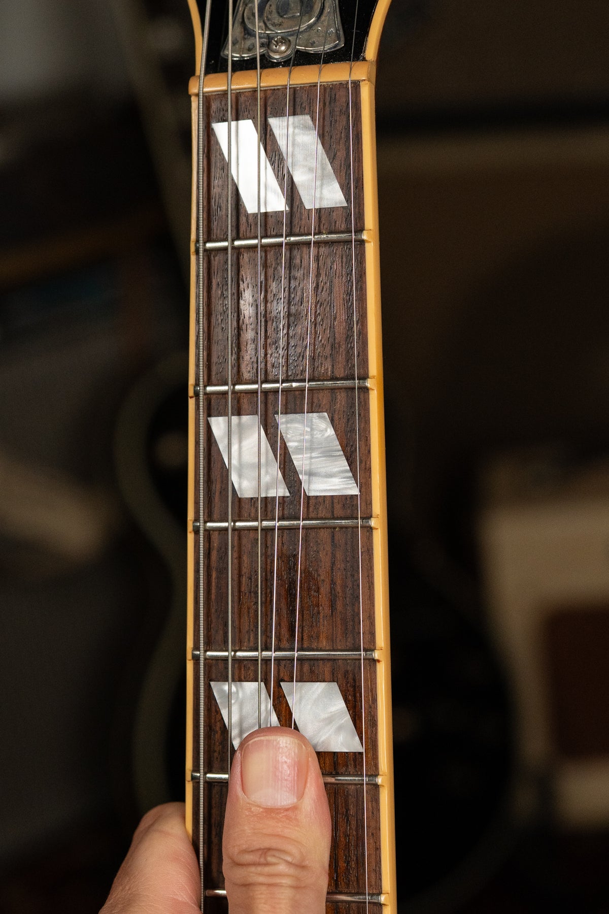 A close-up of a guitar fretboard with trapezoid inlays, a thumb rests on the lowest visible inlay.