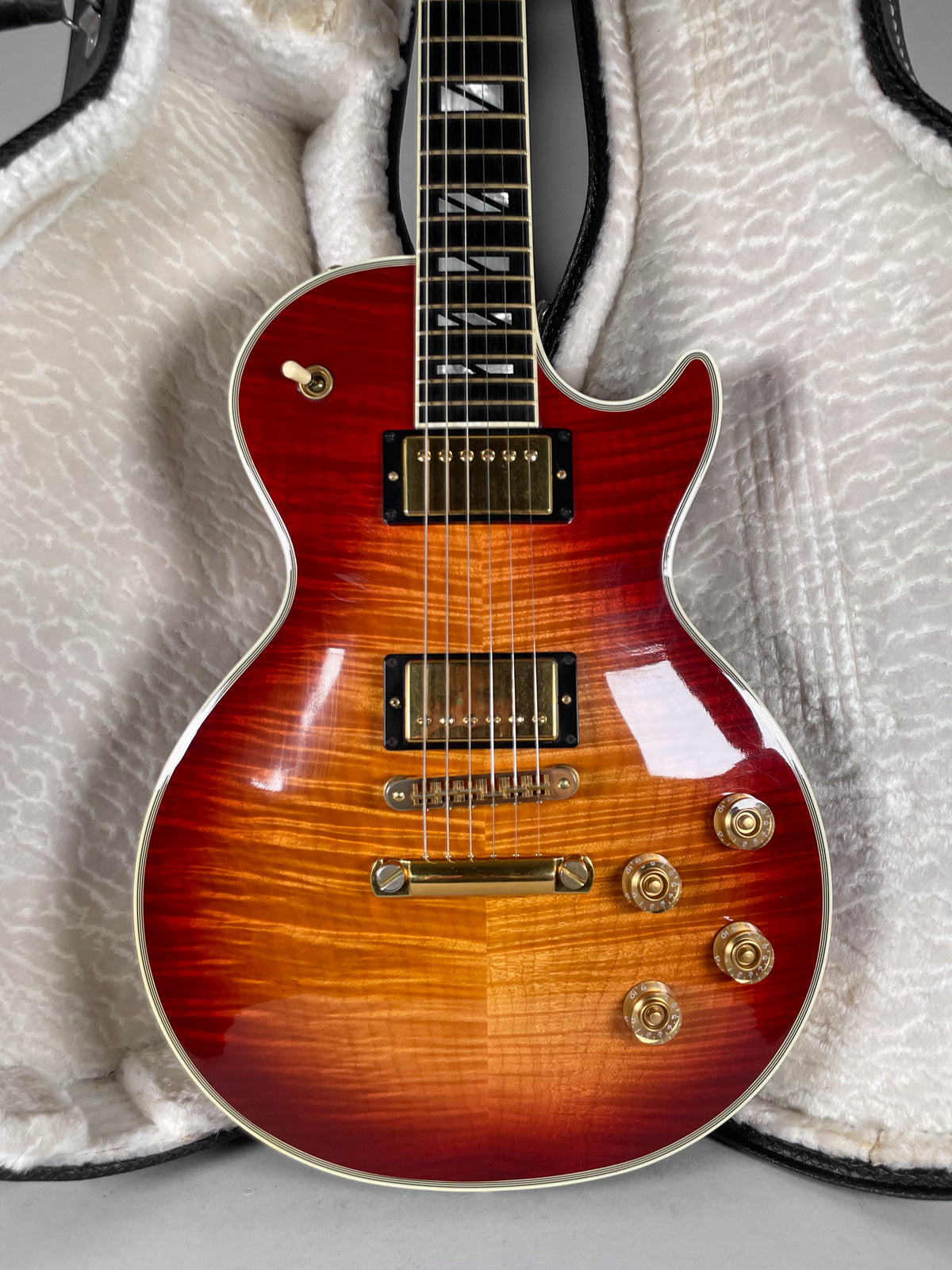 Full-frame view of a cherry sunburst guitar with a flame maple top, gold hardware, and trapezoid inlays, resting in a white plush case.