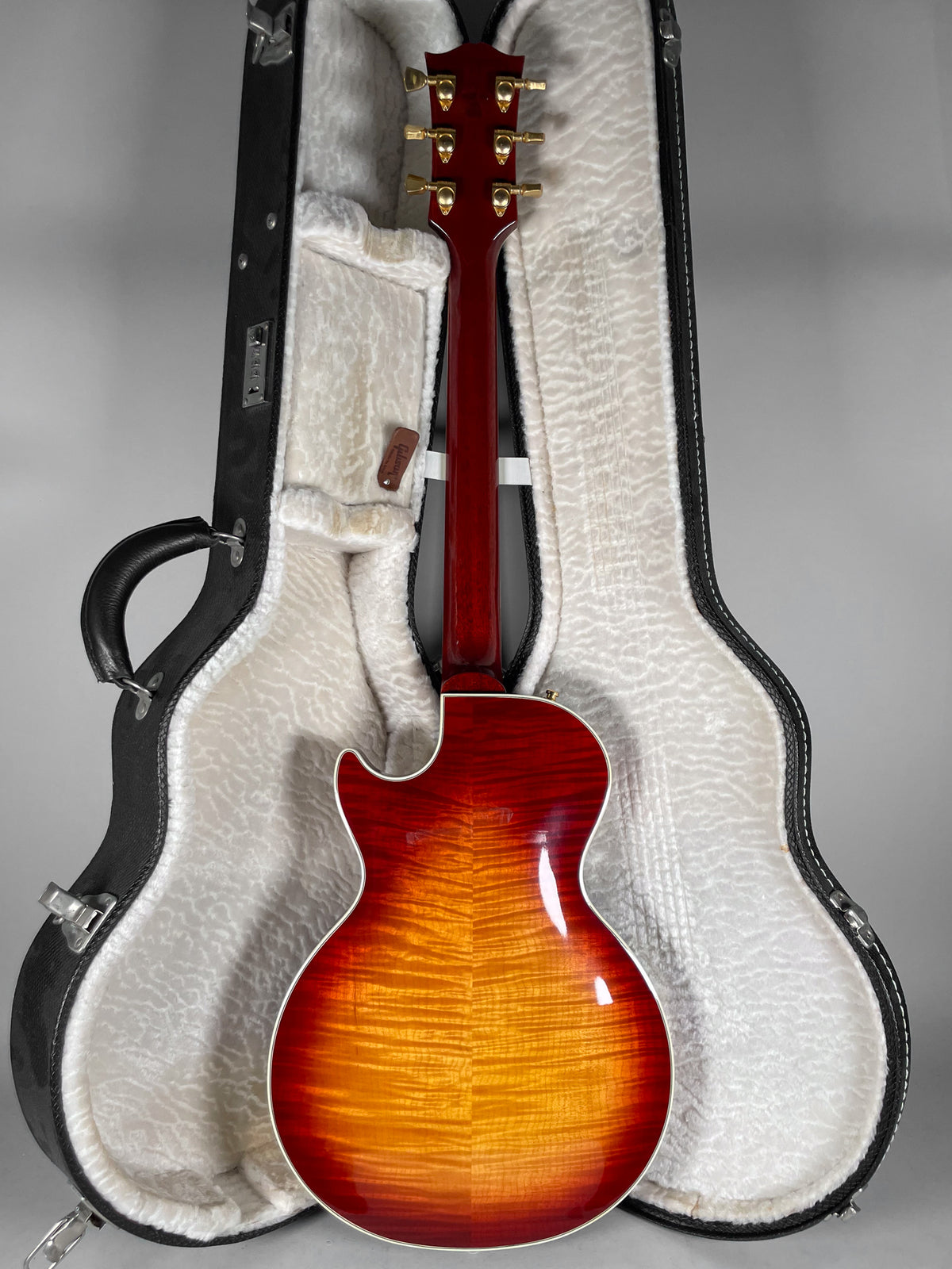 The guitar's cherry sunburst back and neck are visible, with gold tuners, resting in a white plush-lined hard case.