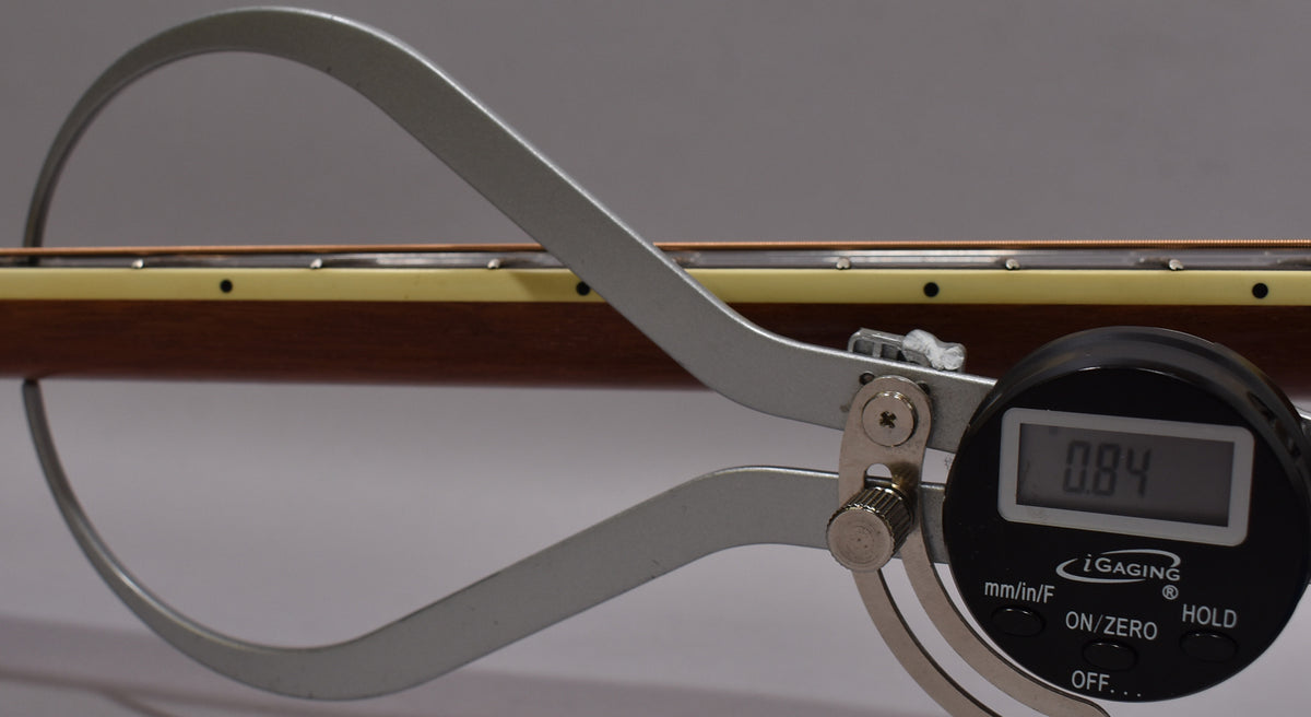A digital caliper measures the neck relief of the natural finish acoustic guitar, showing 0.84 on its display.