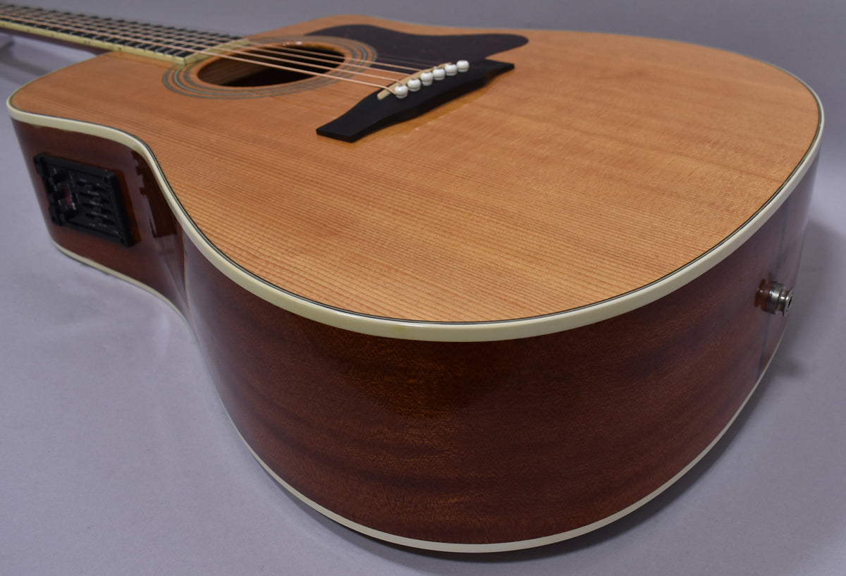 Upper body of a natural finish acoustic guitar, showing the cutaway, preamp controls, and endpin jack.