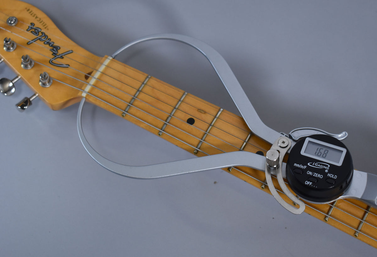Close-up of the guitar's maple neck and headstock, with a digital caliper measuring the string height at the 12th fret, showing 1.68mm.