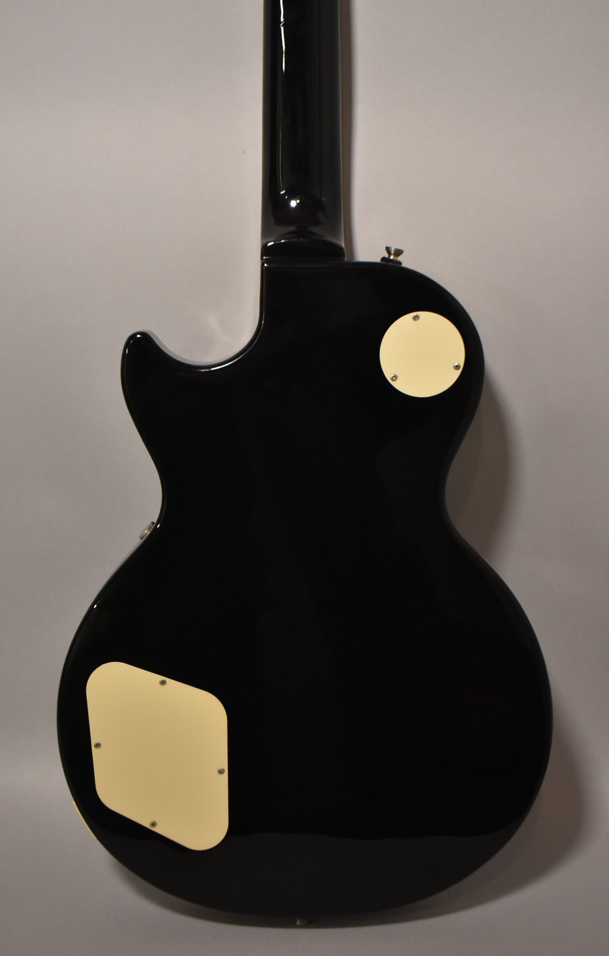 Rear view of a glossy black Les Paul-style guitar, showing the neck and body with two cream-colored control cavity covers.