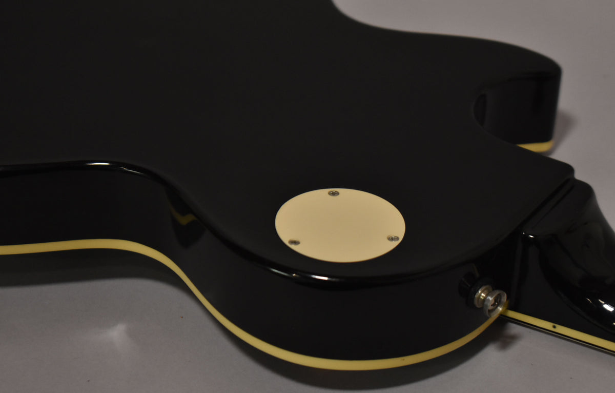 The guitar's glossy black body is shown from the back, revealing a cream-colored control cavity cover and a chrome strap button.