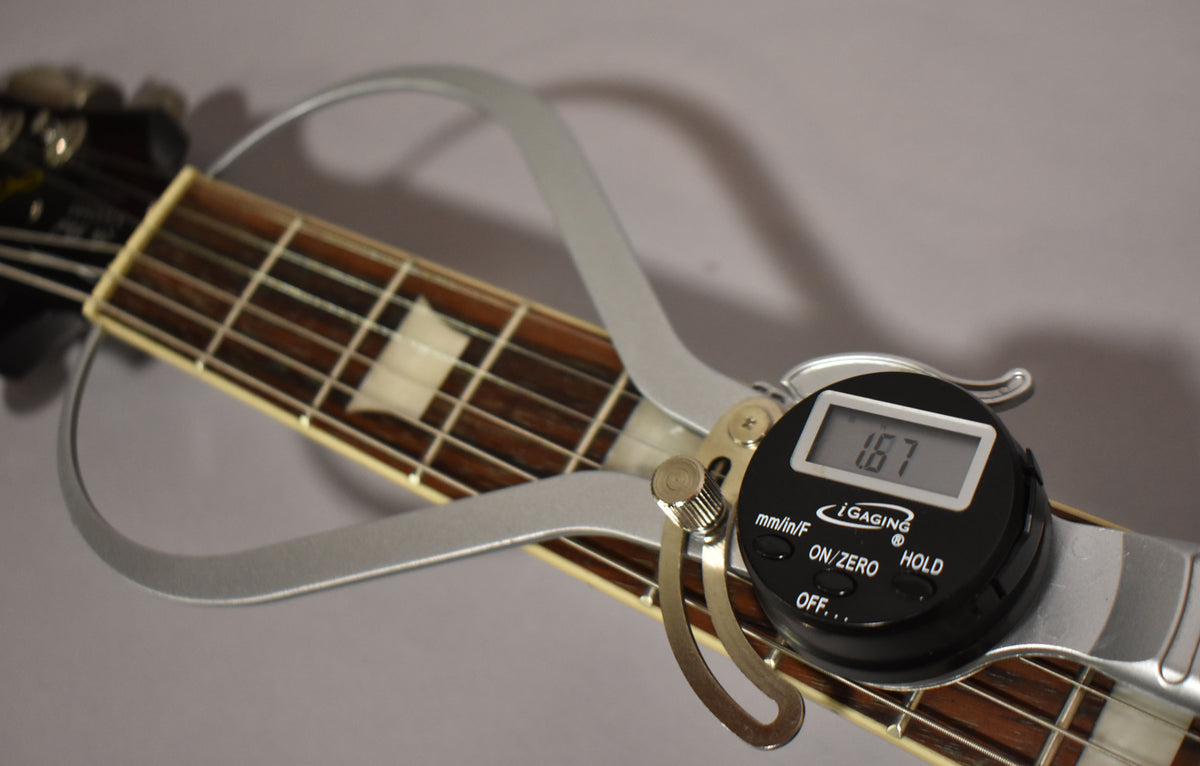 A digital string action gauge shows 1.67mm on the guitar's dark fretboard with trapezoid inlays and cream binding.