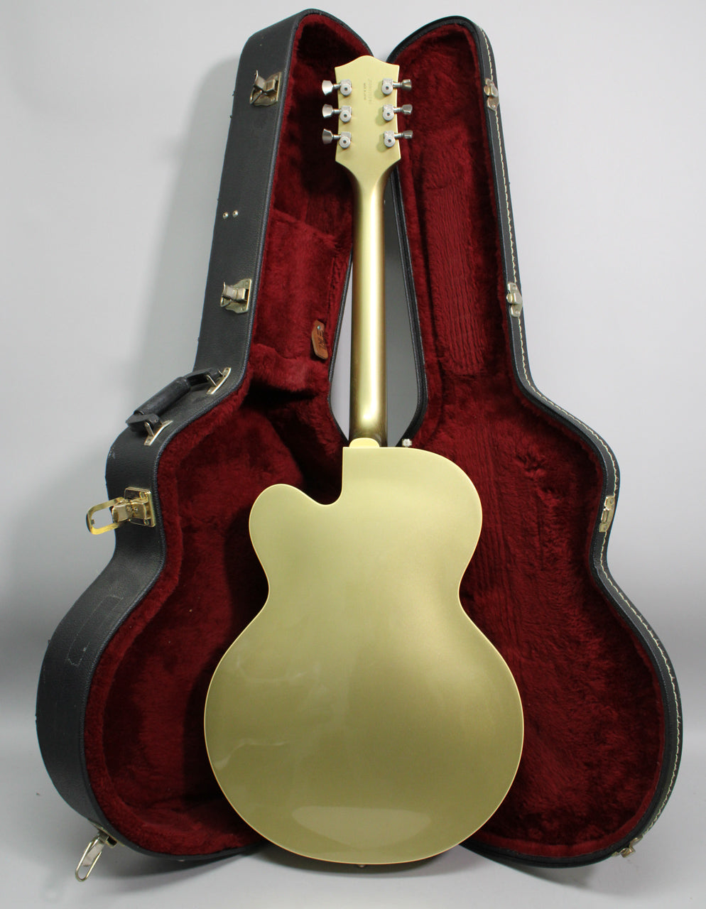 The back of a tan hollow-body guitar with a gold neck and headstock, standing in a black hard case with red plush lining.