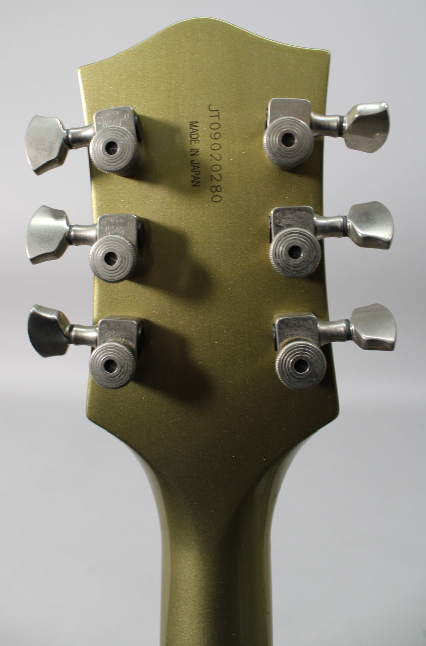 The back of a metallic gold headstock with six brushed chrome tuners, showing light wear and a serial number.