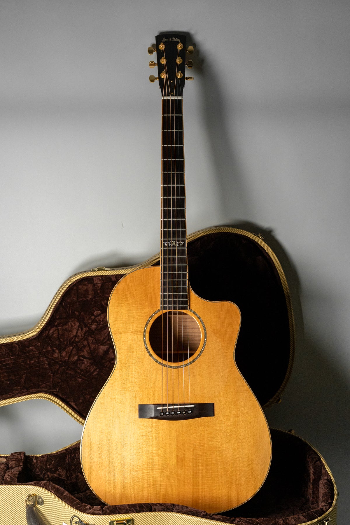 Full-front view of a light-wood acoustic guitar with a cutaway body, dark fretboard, gold tuners, and an open tweed hard case.
