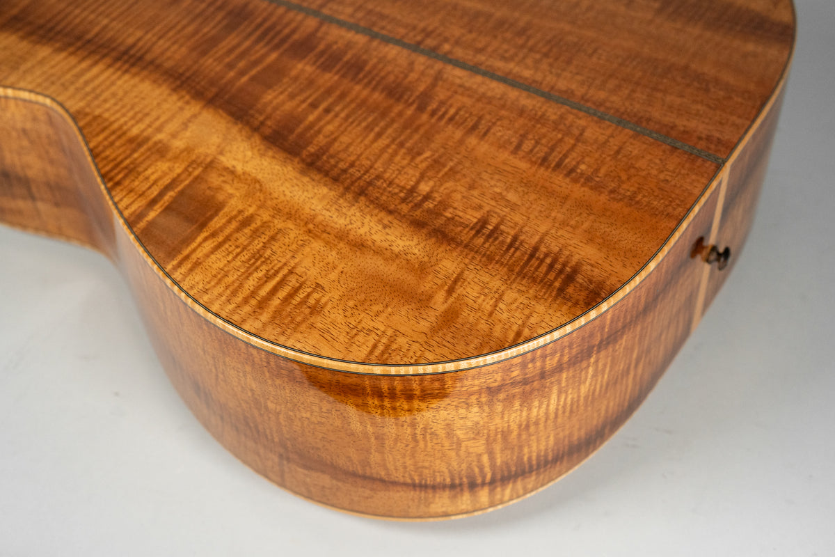 Close-up of the lower bout of a glossy, figured koa acoustic guitar, showing the rich grain and a dark endpin.