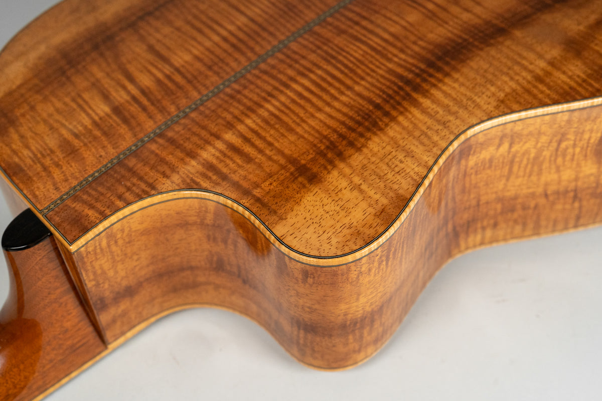 The guitar's glossy, figured koa back and side are visible from the upper bout to the waist, with a decorative backstrip.