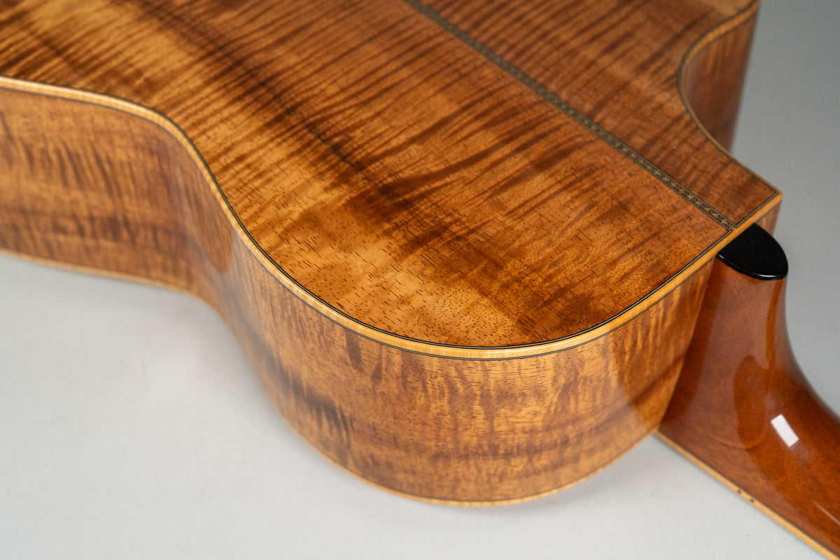 The guitar's lower bout and cutaway are shown from the back, revealing highly figured koa wood with a dark center strip.