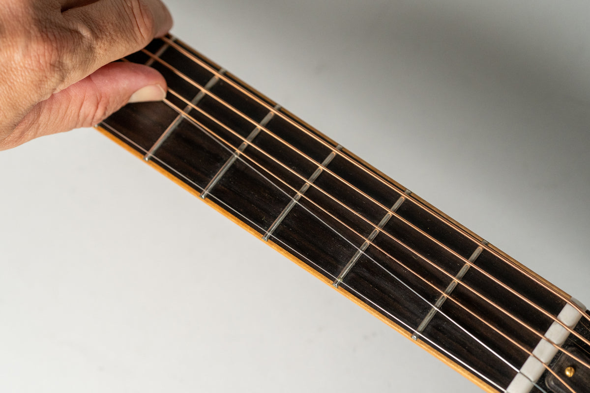 A hand frets the dark rosewood fingerboard of the acoustic guitar, showing the first five frets and copper-colored strings.