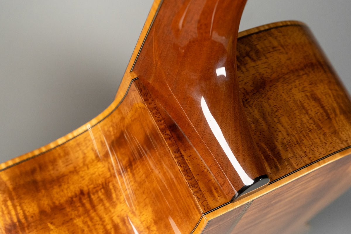 Close-up of the guitar's glossy, figured koa body and neck heel, showing light surface scratches on the lower bout.