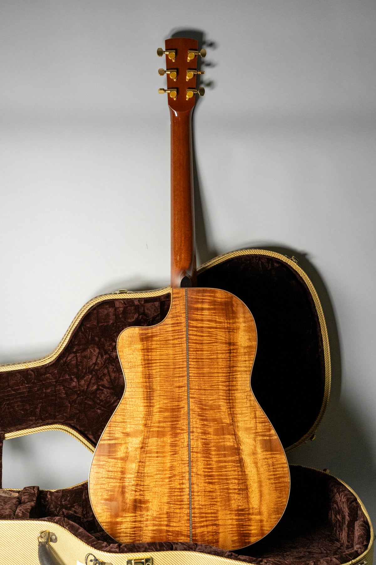 Full back view of a glossy, amber-orange flamed koa guitar with a cutaway, dark brown neck, gold tuners, and an open tweed case.