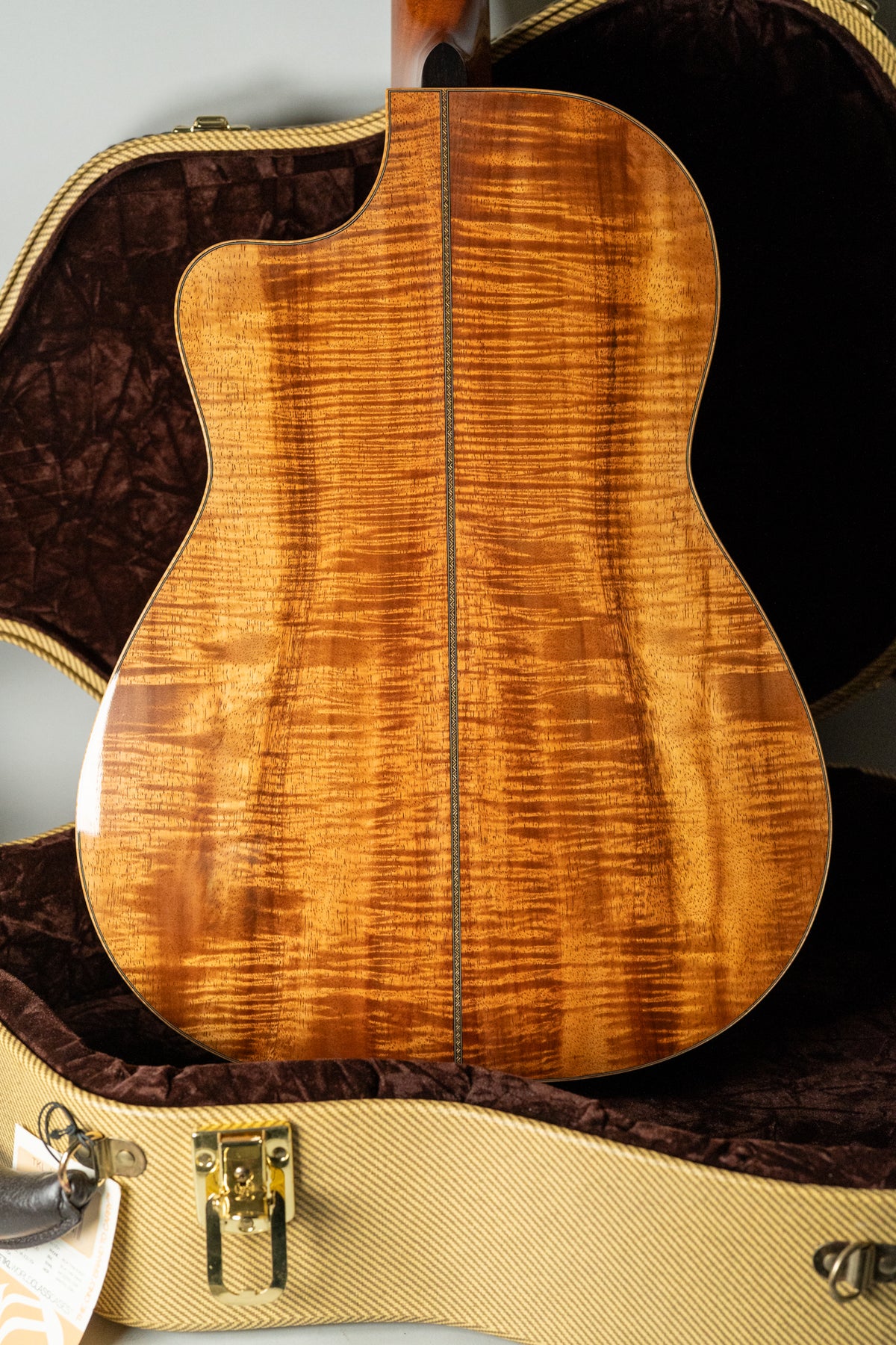 The guitar's highly figured koa back, with a central seam, is visible from the upper bout to the lower bout, resting in its case.
