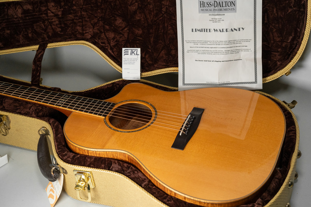 The light brown acoustic guitar, with a cutaway body and dark bridge, rests in a tweed case with a brown velvet interior.