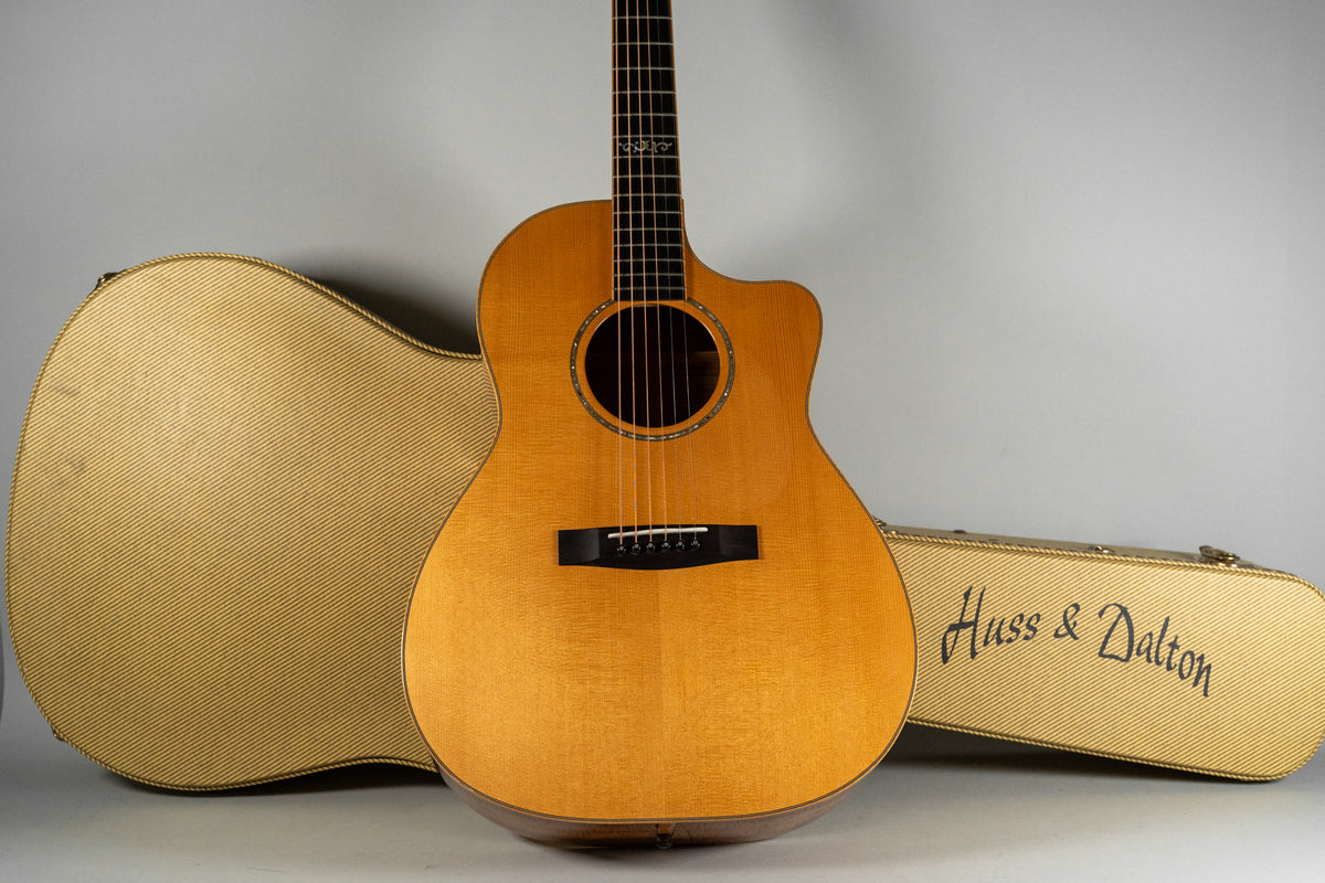 Light brown acoustic guitar with a cutaway, dark bridge, and ornate fretboard inlays, flanked by two tweed cases.