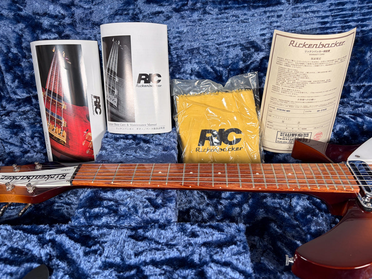 The guitar's dark brown body and neck are visible, with two manuals, a warranty card, and a yellow cleaning cloth in a blue velvet case.