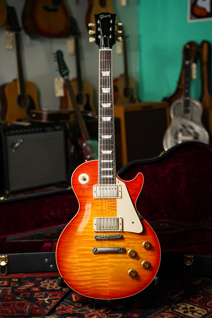 Full-body shot of a sunburst electric guitar with a flamed maple top, cream binding, and gold hardware, standing in its open case.