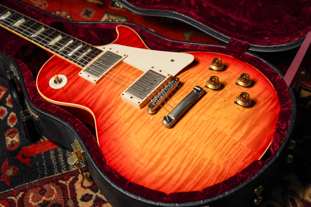 The sunburst electric guitar, with a flame maple top and cream binding, rests in its open, red velvet-lined hard case.