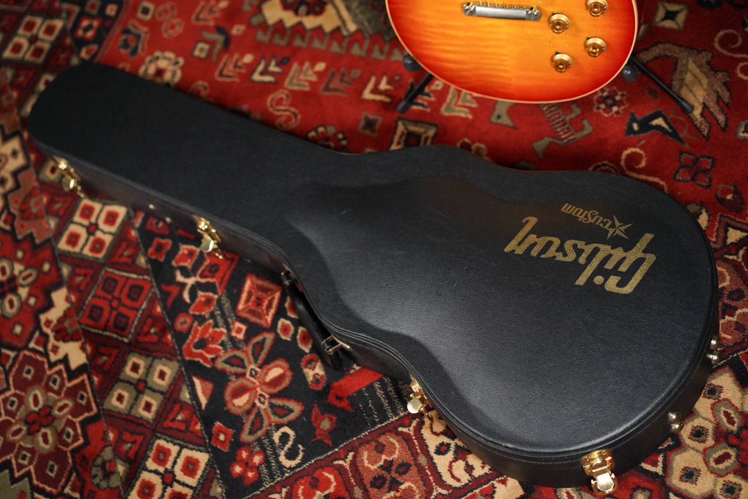 A black hard case with gold latches and a gold logo is angled on a patterned red rug, with a sunburst guitar body visible at the top.