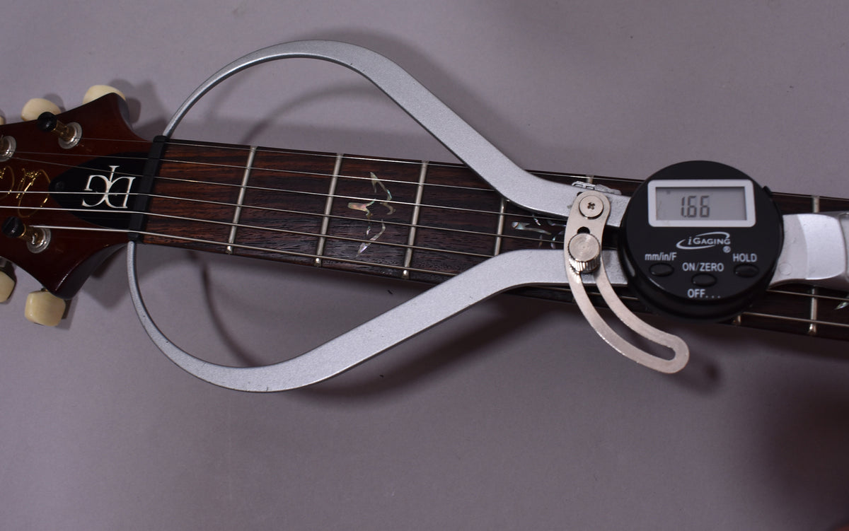 A digital caliper measures the dark rosewood fretboard, showing 1.66 on the display, with bird inlays and cream tuning pegs visible.