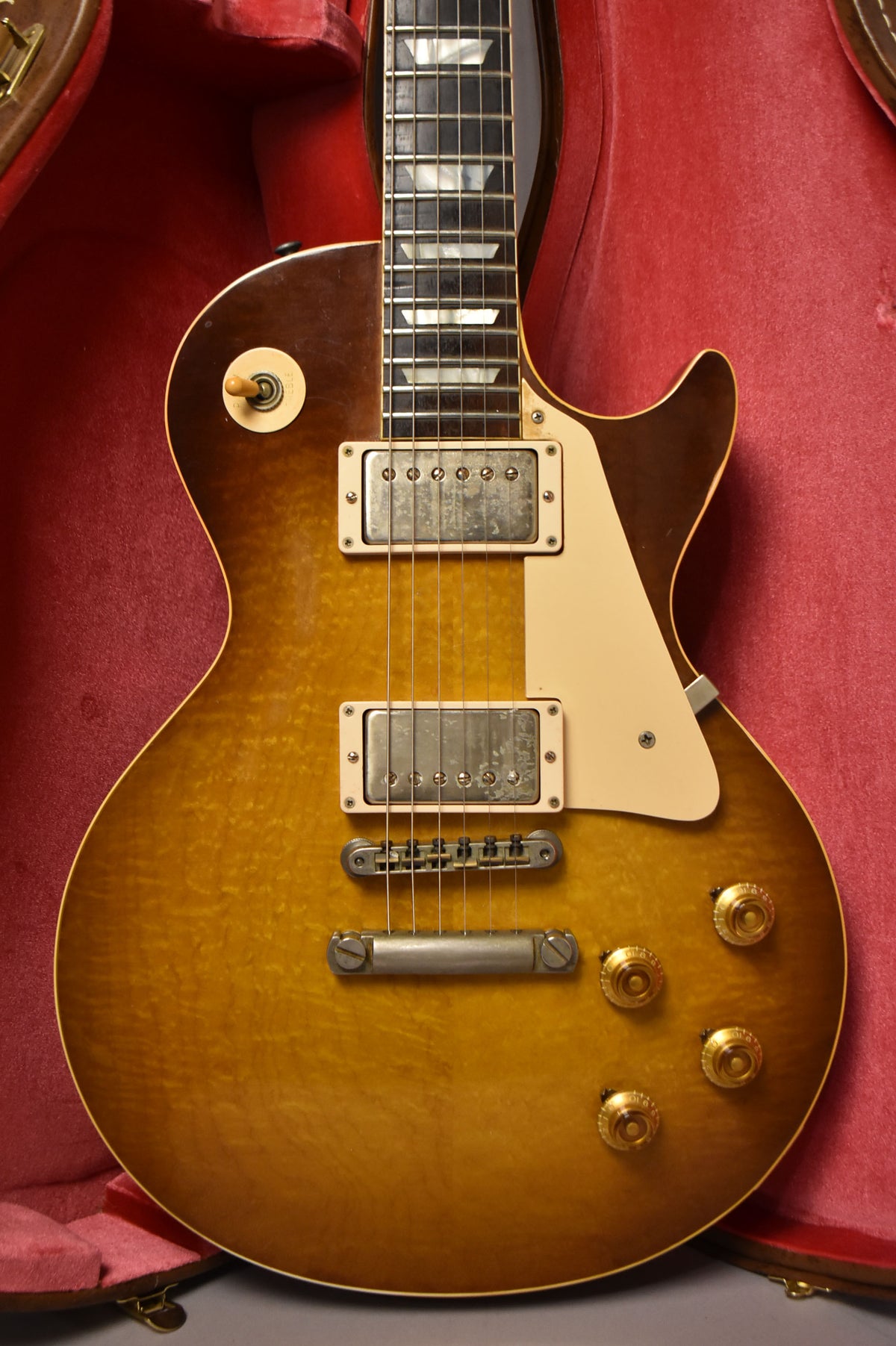 The guitar's sunburst body, with visible wood grain, is framed in a red case, showing two humbuckers and four gold knobs.
