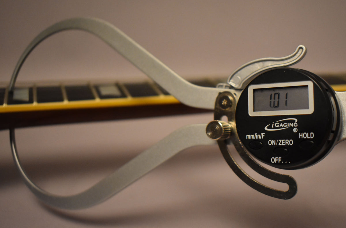 A digital caliper measures the guitar's fretboard, showing "1.01" on its display, with block inlays visible.