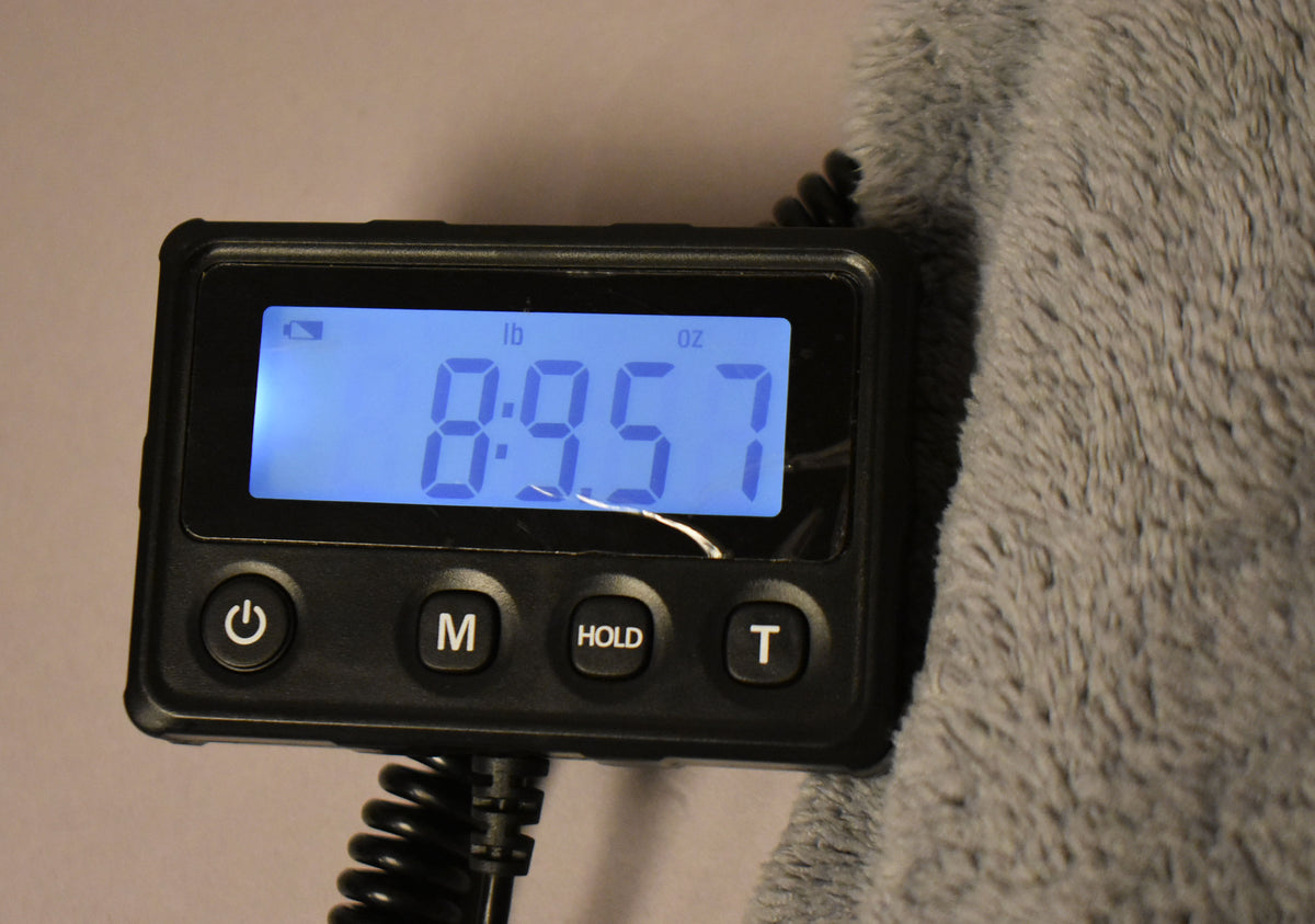 A digital scale displays "8:9.7" in blue, indicating 8 pounds, 9.7 ounces, next to a gray furry object.
