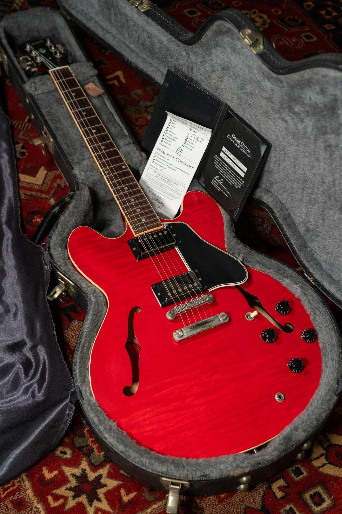Cherry red flamed maple guitar in its open hard case, angled from headstock to body, with paperwork on the body.
